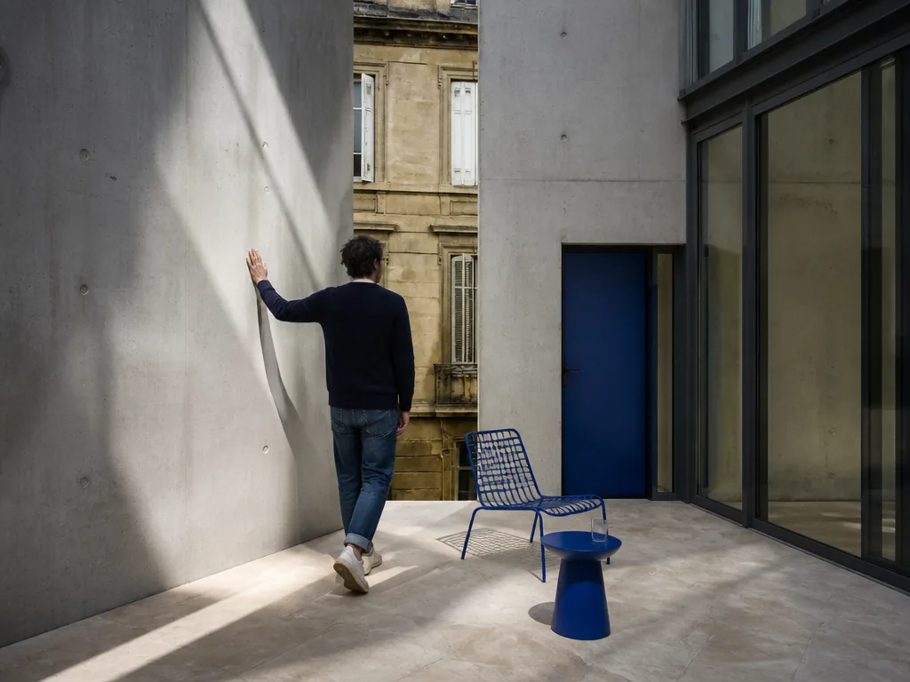 Photorealistic editorial photograph of a resident touching a raw concrete wall in a new Bordeaux courtyard, sun casting 